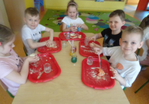 Dzieci mieszają składniki na ciastolinę na tackach rozłożonych przed każdym dzieckiem na stole, jeden chłopiec barwi masę na zielono.